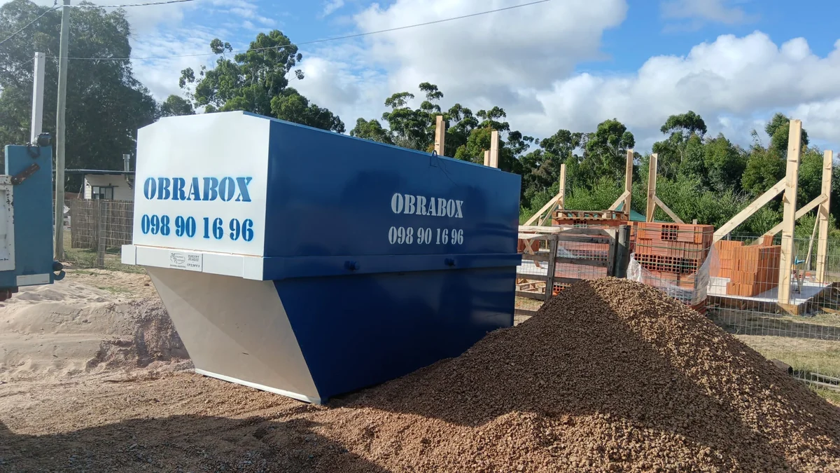 Obrador ObraBox instalado junto a un acopio de arena, con la obra en construcción al fondo